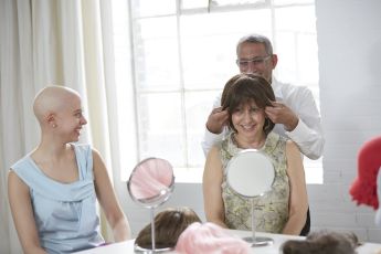 Wig stylist placing human hair wig on female cancer survivor as another female cancer survivor watches