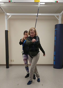 Image of a woman taking a step while wearing a harness attached to a metal structure; a physiotherapist stands behind the woman.